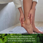 A person standing on a gray rug, bent over with hands touching one foot, following a foot care routine. Text on the image reads, "Epsom Salt Foot Soak with Tea Tree Oil naturally rids the skin of the causes of smelly feet." Green leaves are visible at the bottom.