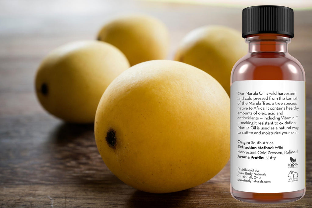 A bottle of Marula Facial Oil with product information on the label is in the foreground. In the background, there are four yellow marula fruits on a wooden surface. The label details the oil's origin, extraction method, and aroma profile.
