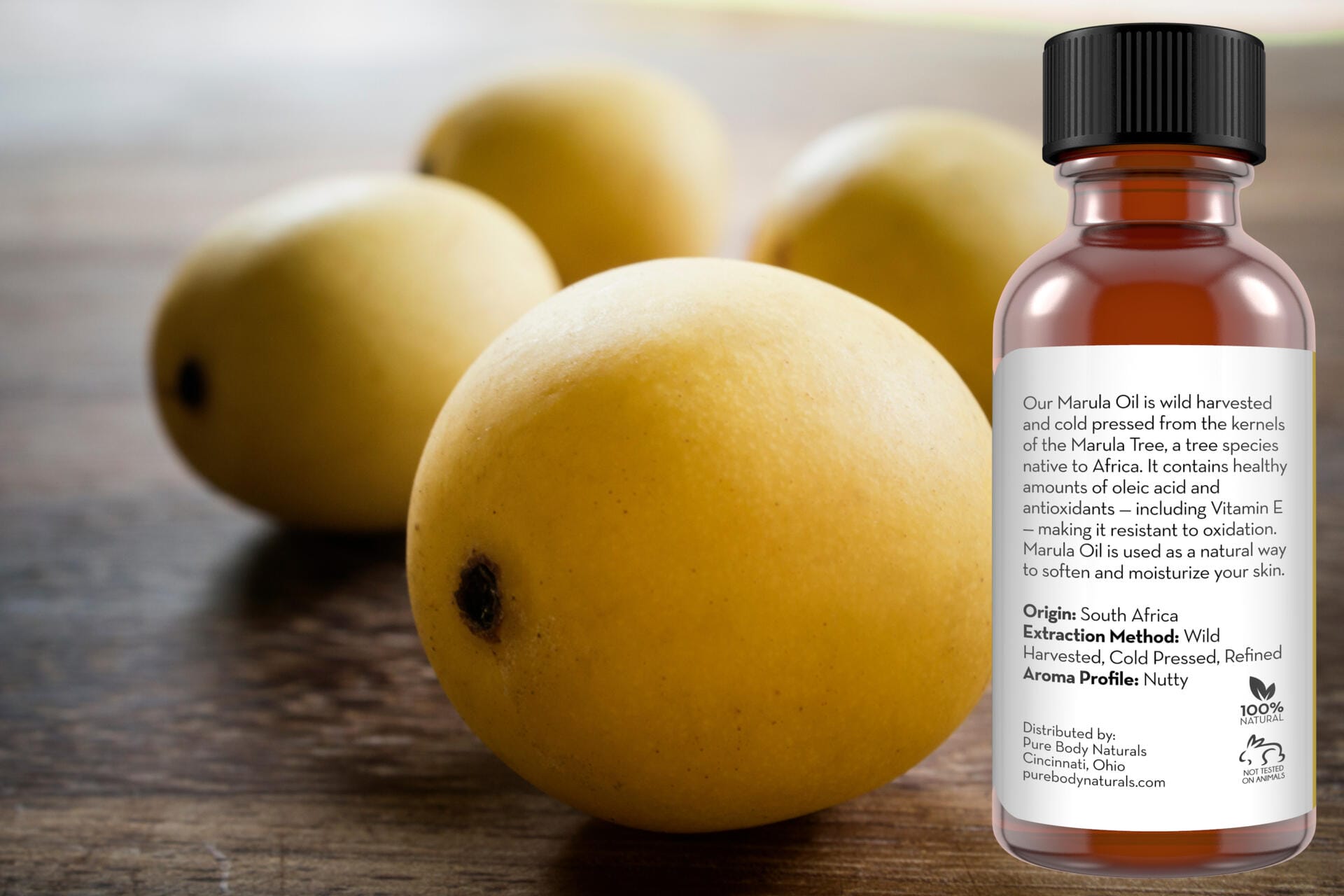 A bottle of Marula Facial Oil with product information on the label is in the foreground. In the background, there are four yellow marula fruits on a wooden surface. The label details the oil's origin, extraction method, and aroma profile.