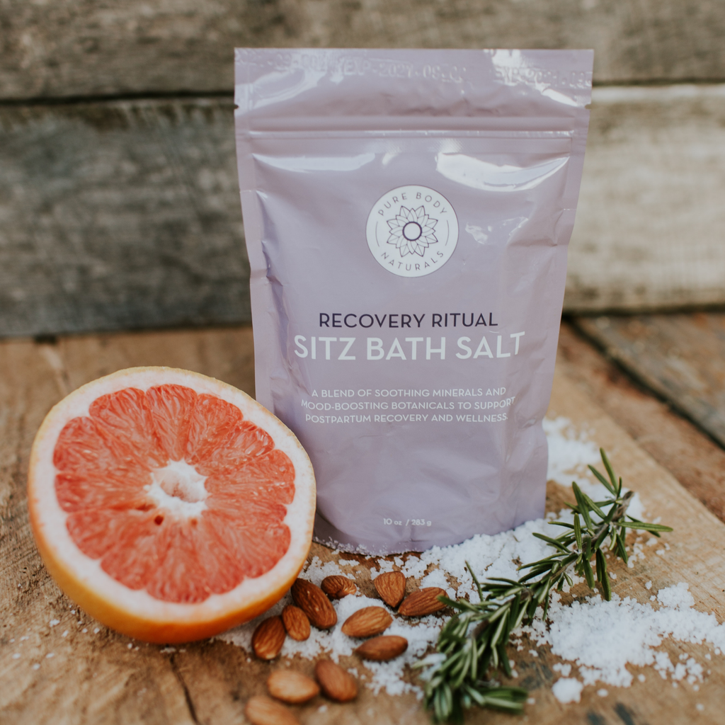 A packet of "Recovery Ritual Sitz Bath Salt, 10 oz" is surrounded by a halved grapefruit, almonds, rosemary sprigs, and scattered sea salt. The packet is lavender in color, placed on a rustic wooden background.