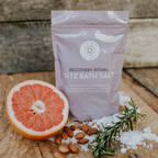 A packet of "Recovery Ritual Sitz Bath Salt, 10 oz" is surrounded by a halved grapefruit, almonds, rosemary sprigs, and scattered sea salt. The packet is lavender in color, placed on a rustic wooden background.