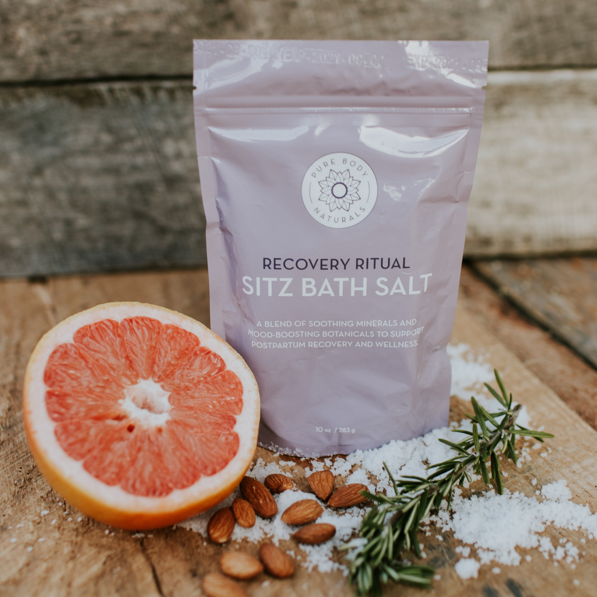 A packet of "Recovery Ritual Sitz Bath Salt, 10 oz" is surrounded by a halved grapefruit, almonds, rosemary sprigs, and scattered sea salt. The packet is lavender in color, placed on a rustic wooden background.